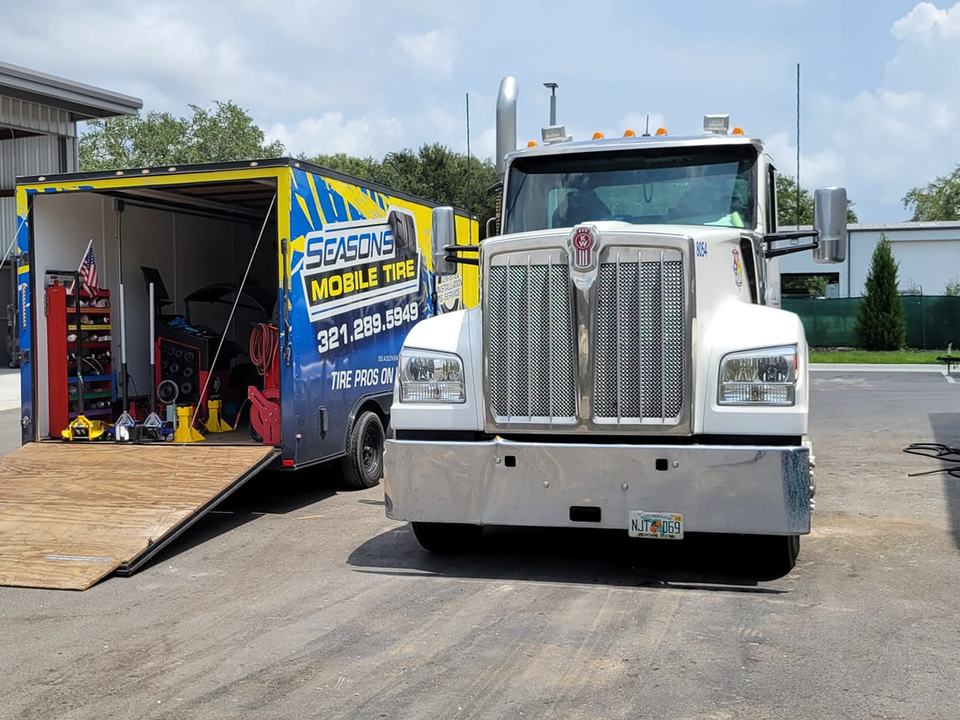 A white Kenworth semi parked nose-out beside the open Seasons Mobile Tire trailer