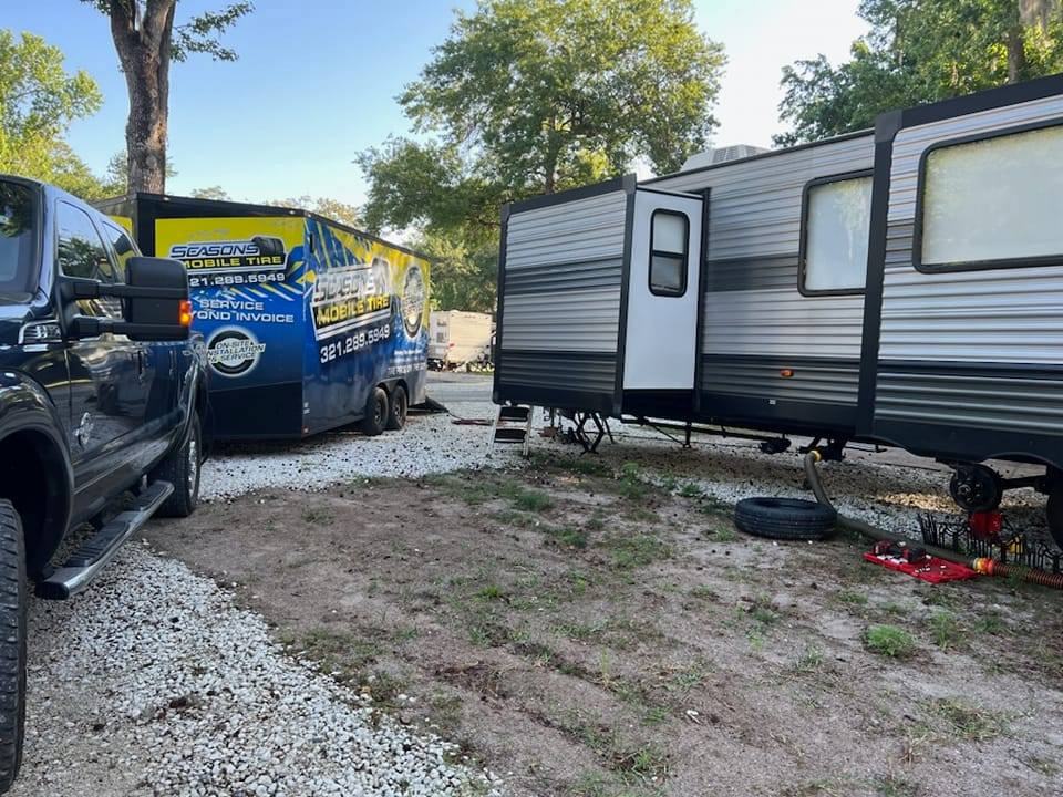 A travel trailer jacked up on a gravel lot with the Seasons trailer and a pickup beside it